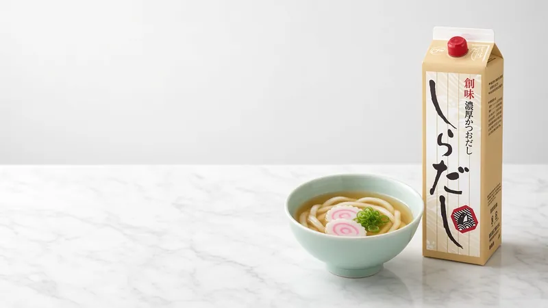 Clear golden udon broth with narutomaki fish cake and spring onion in a celadon bowl beside a Somi shiradashi 1.8 litre carton